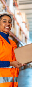 Professional female warehouse worker in hard hat and safety vest holding a carton, smiling confidently amid busy distribution center aisles stacked with packages and inventory